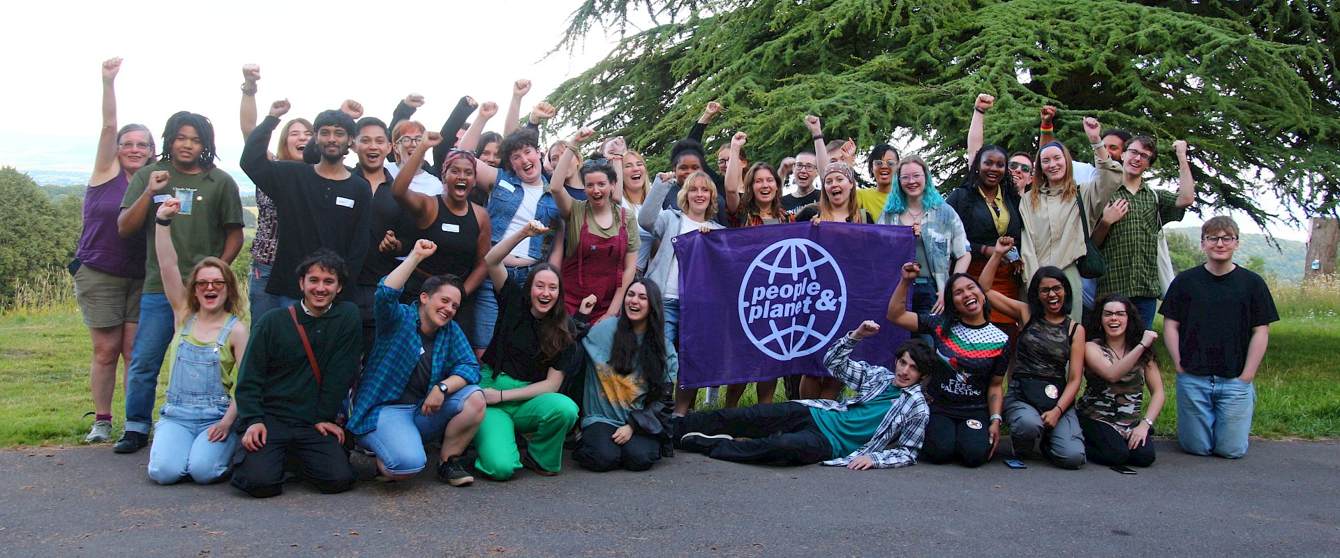 Photo of large group of students holding up a People & Planet banner with fists in the air outdoors by large tree