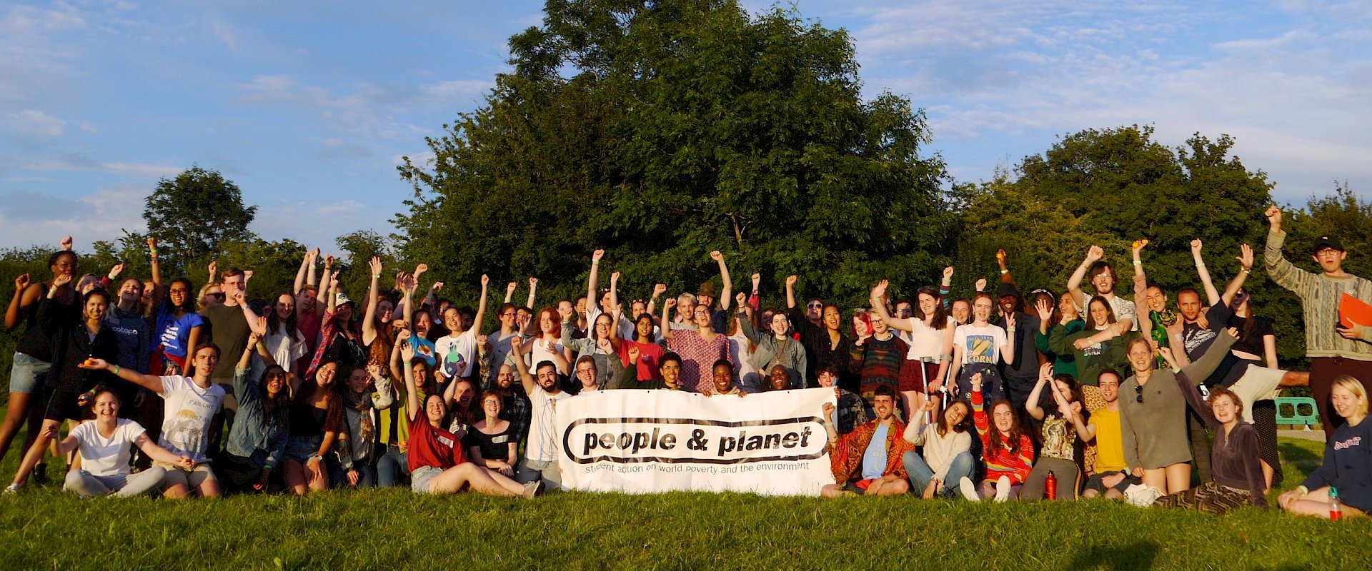 Photo of a hundred or so young people holding fists in the air at an outdoors training event called Power Shift, behind a People & Planet banner.