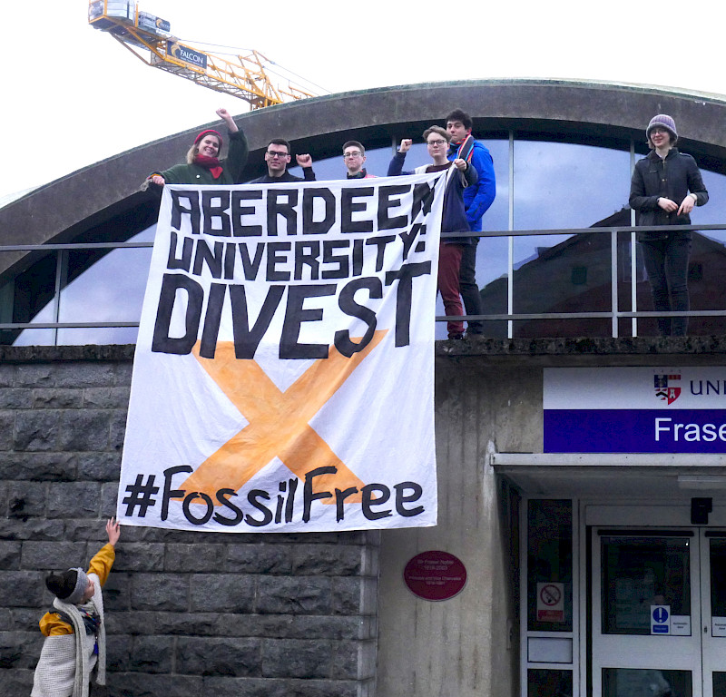Photo of 6 students on top of a building displaying a massive banner reading: Aberdeen university DIVEST #FossilFree