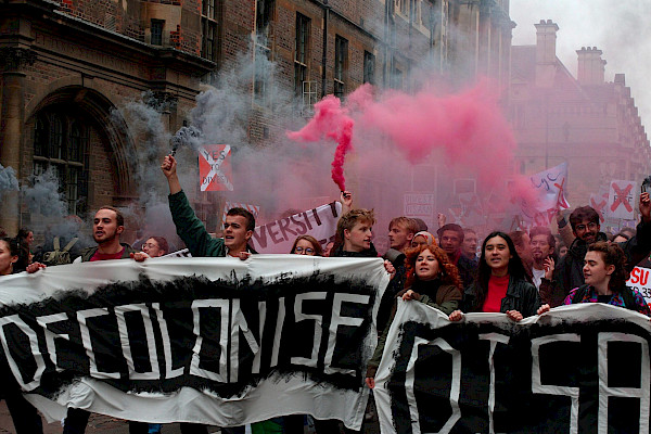 Photo of a demonstration with pink smoke and people marching behind a massive banner reading: Decolonise DISA. Over the image is the Divest Borders campaign logo.
