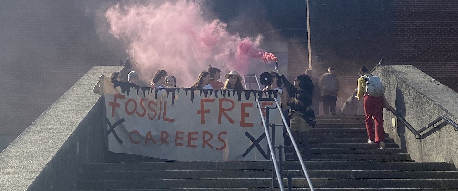 Image of students standing on steps outside of a univeristy building and holding a Fossil Free Careers banner. The students are also holding red smoke flares.
