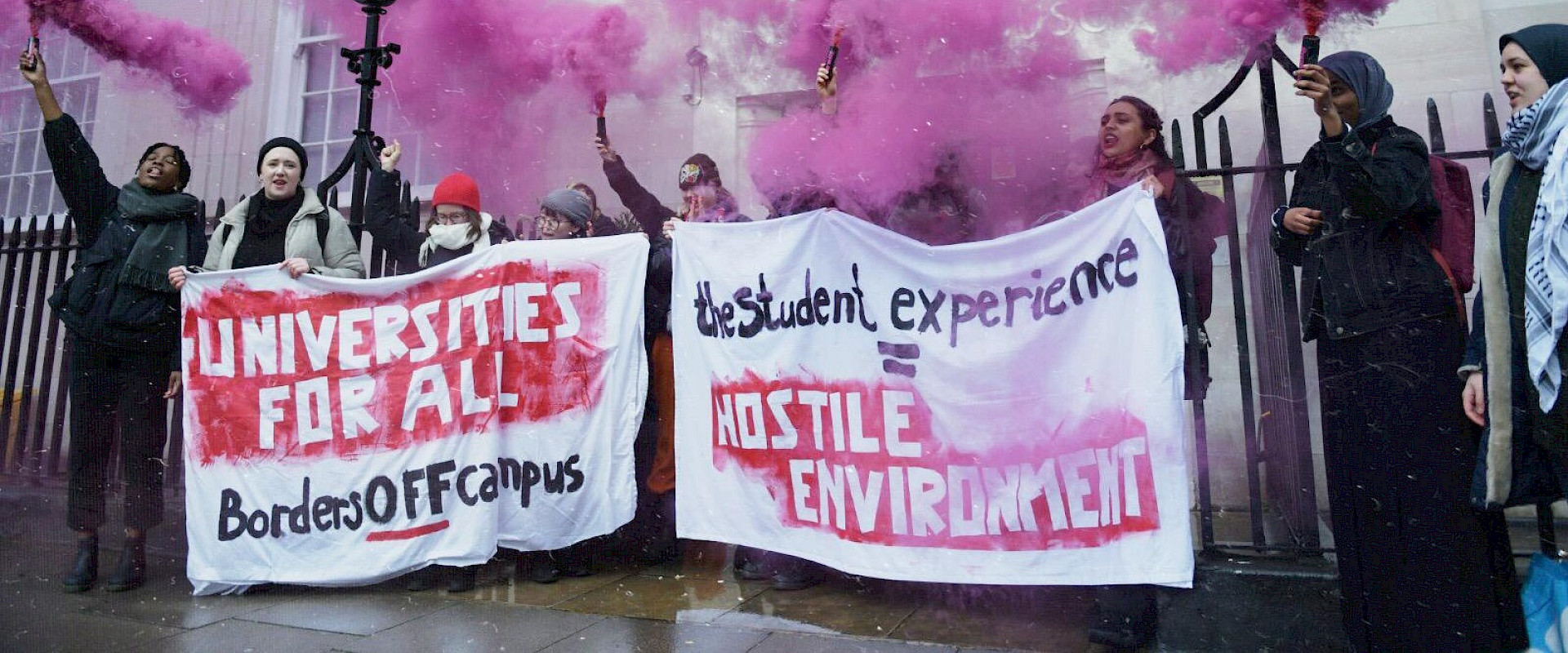Crowd of students holding up banners reading "Universities for all: Borders Off Campus: Hostile Environment" with pink flares