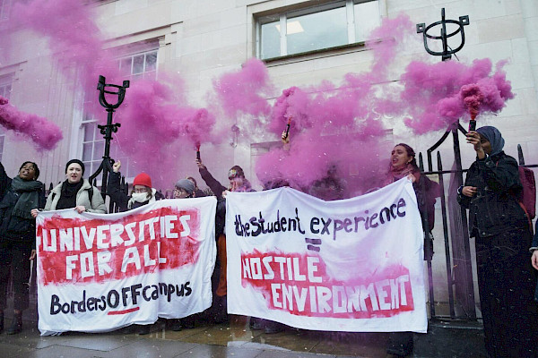 Crowd of students holding up banners reading "Universities for all: Borders Off Campus: Hostile Environment" with pink flares