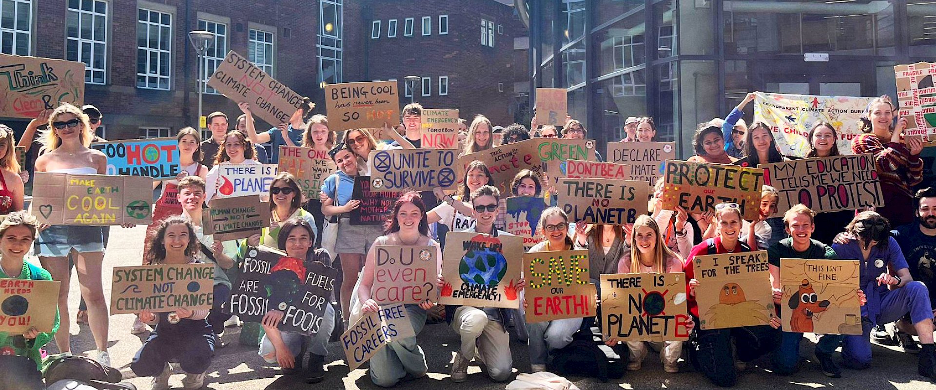 Large group of students holding up placards in the sunshine outdoors. Placards read phrases such as "there is no planet b", and "system change, not climate change"