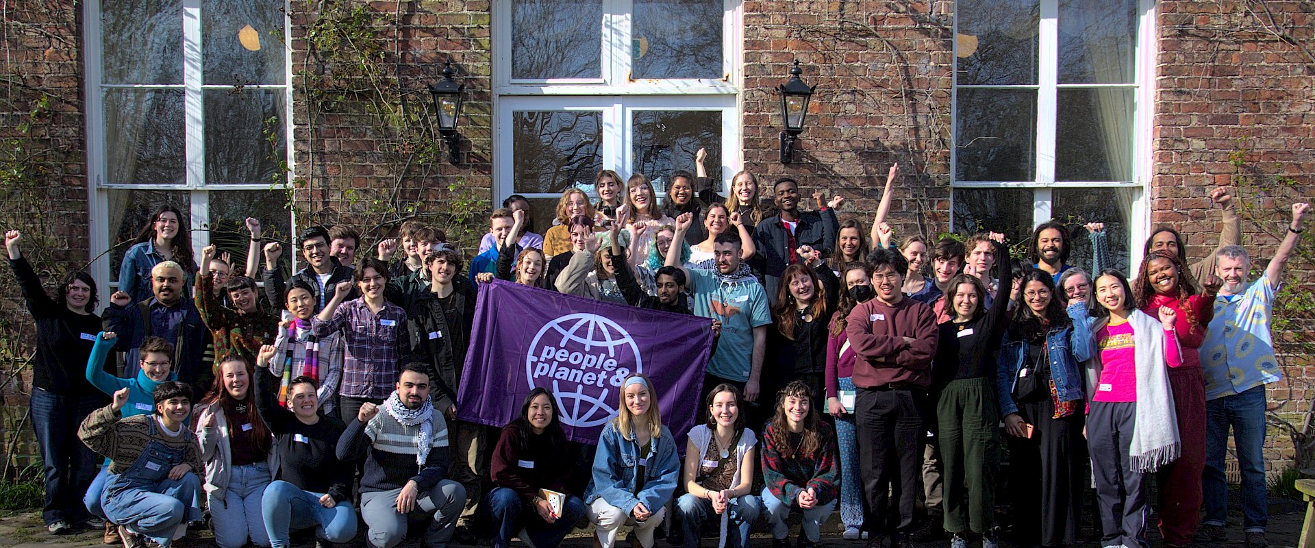Large group of students with people & planet flag raising their arms and smiling in front of a building