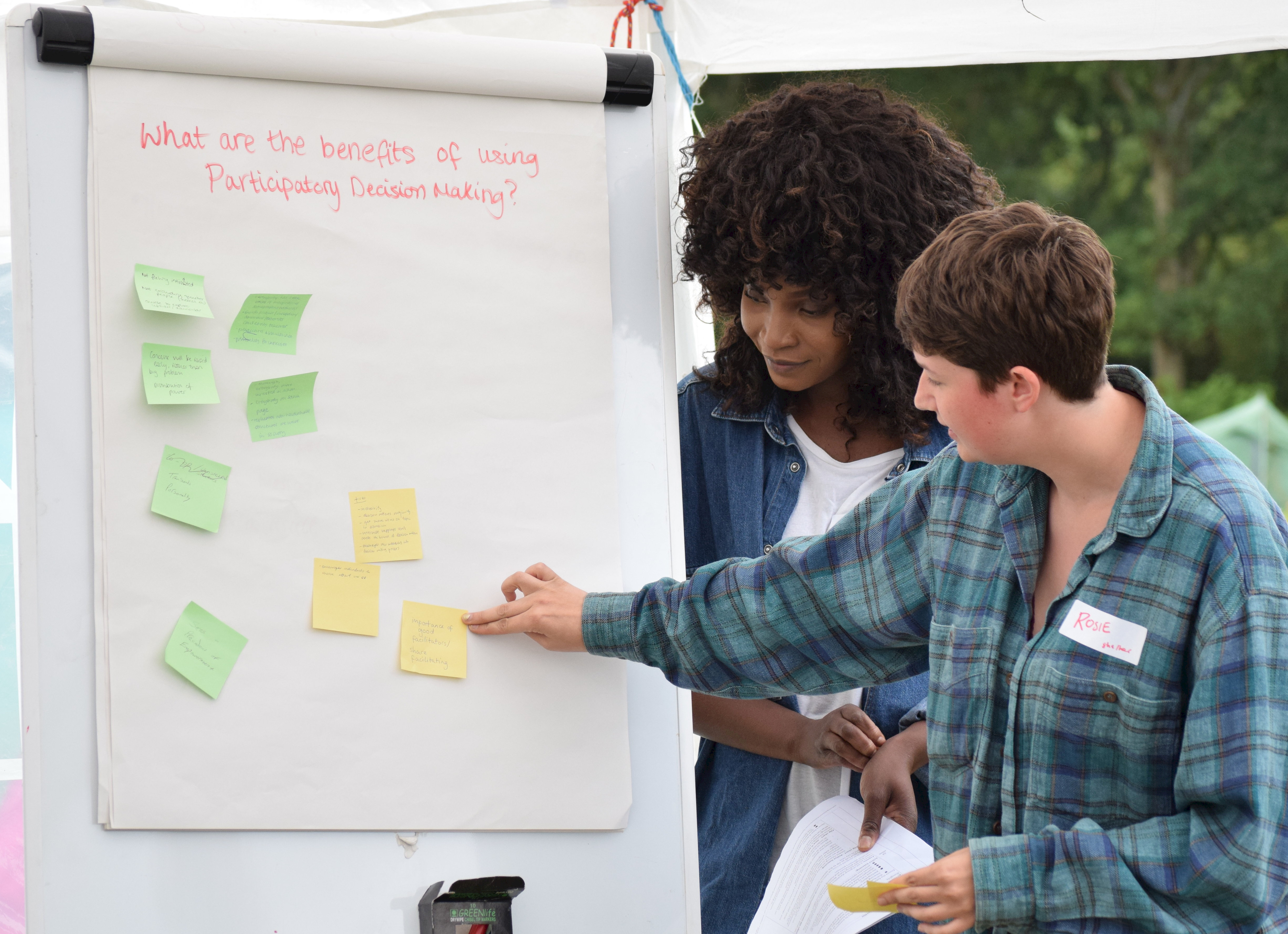 Two young campaigners leading a workshop stand in front of a flipchart paper which says 'what are the benefits of participatory decision making'?'