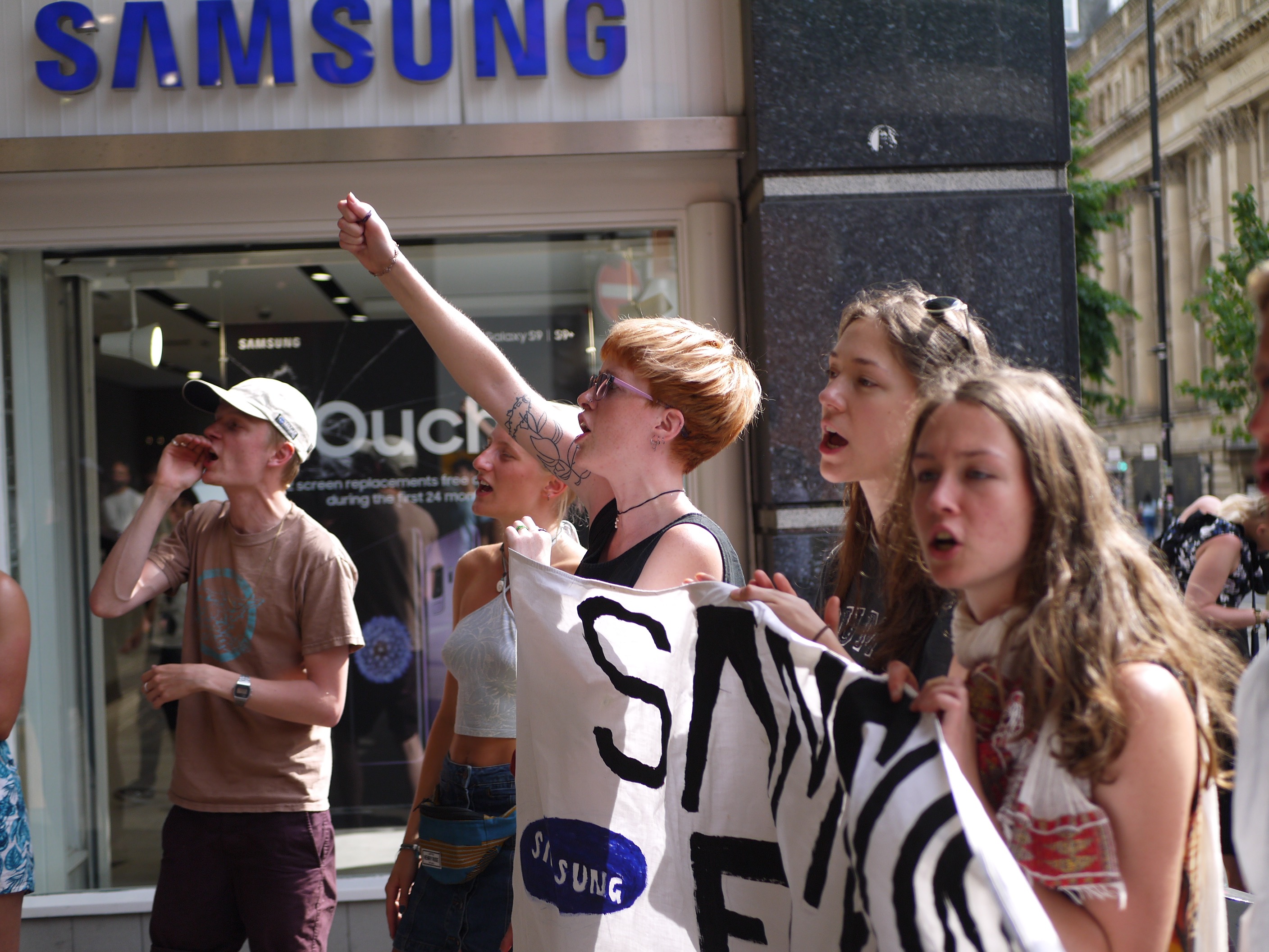 five student activists protest outside a Samsung outlet. They are holding a banner and chanting.