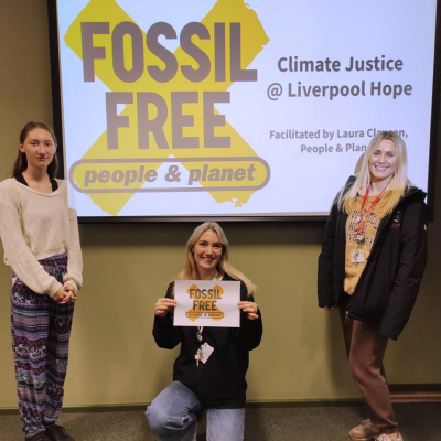 Three students from Liverpool Hope University stand in front of a P&P Fossil Free workshop slide, one is holding a poster with the Fossil Free logo