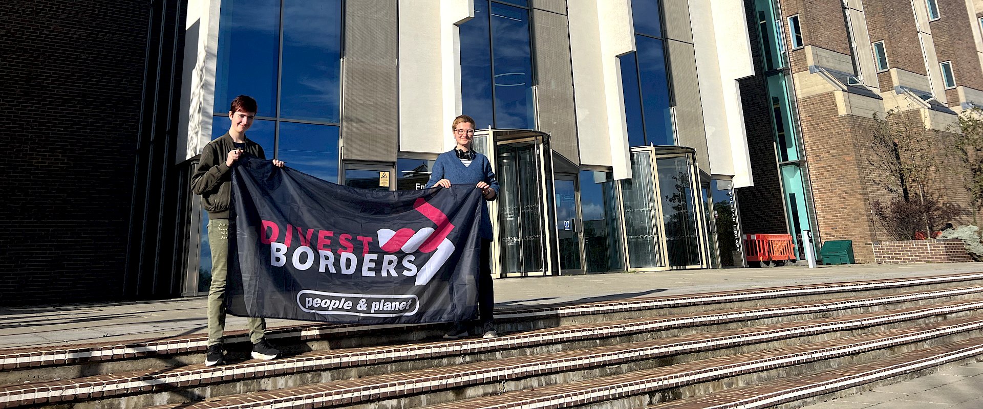 Two students stand with Divest Borders banner in front of university