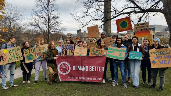 Crowd of students stand outside holding up handmade placards with climate justice slogans on and a banner that reads 