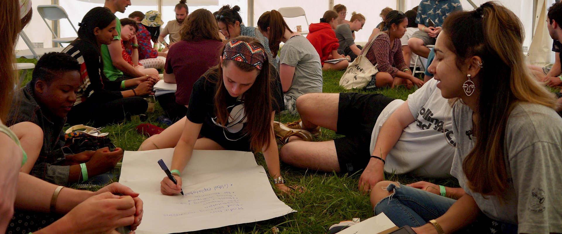 Students sitting in groups on grass in large marquee writing on flip chart paper