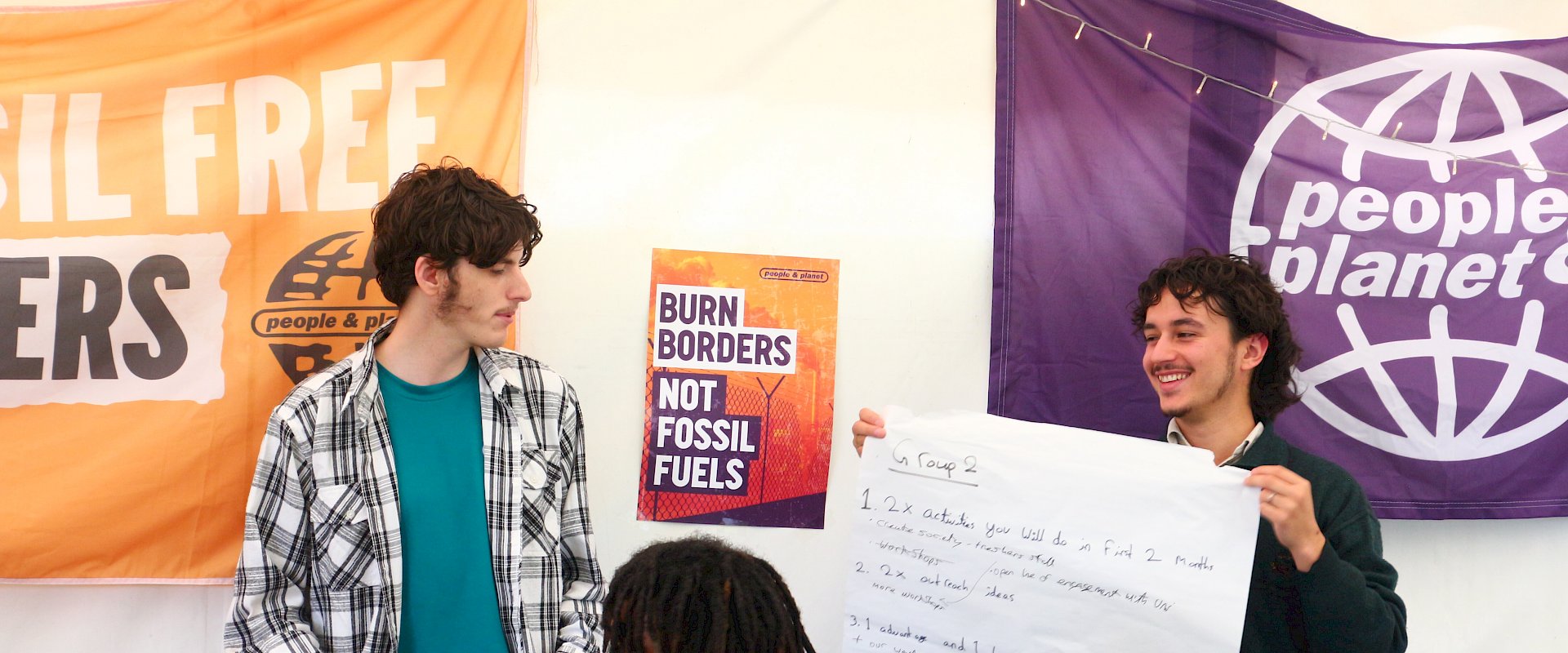Coordinator and student stood holding up flipchart paper with some notes on. Banners and posters are on the walls that say People & Planet, "Burn borders, not fossil fuels".